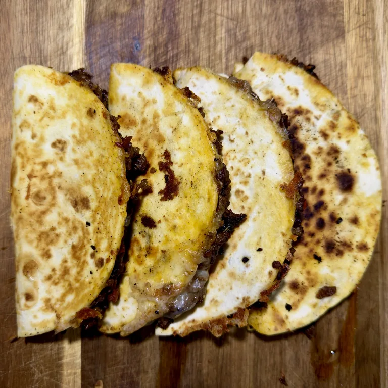 Four folded browned sausage and Oaxaca cheese tacos on a wooden cutting board