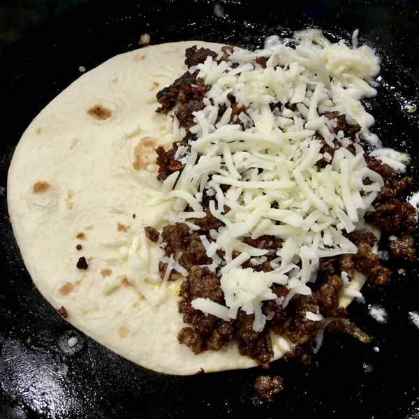 Sausage filling and shredded Oaxaca cheese on one half of an open flour tortilla in a skillet