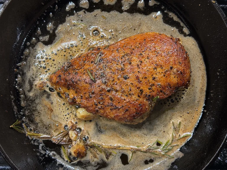 Finished seared chicken breast in a cast iron skillet surrounded by foaming garlic and rosemary butter